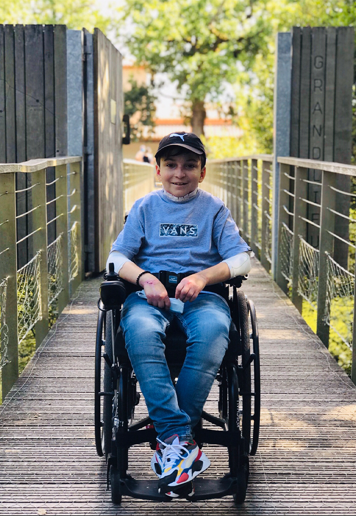 A smiling boy in a wheelchair on a wooden ramp with metal barriers and trees in the background.