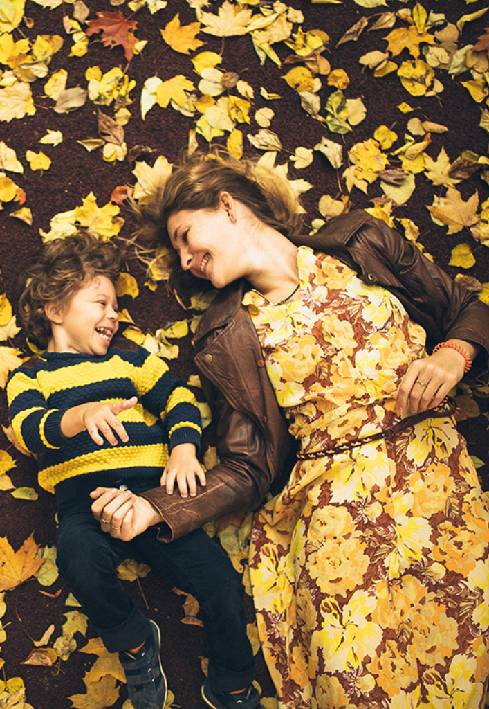 A mother and her child laugh while lying on their backs, surrounded by autumn leaves and dressed in clothes that match the yellow and brown colours of the scene.