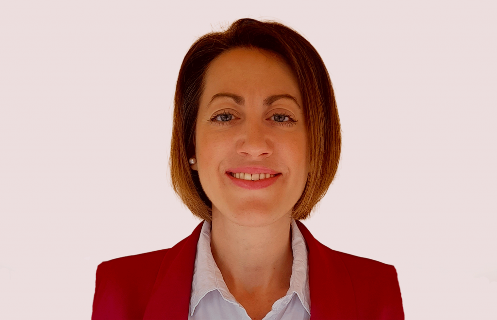 Headshot of Anna Spinou. She wears a red jacket and smiles at the camera.