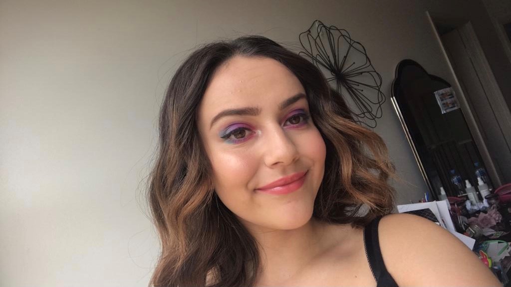 Angelina smiles at the camera as she takes a selfie in her bedroom. She has bold blue, purple and pink eyeshadow in.