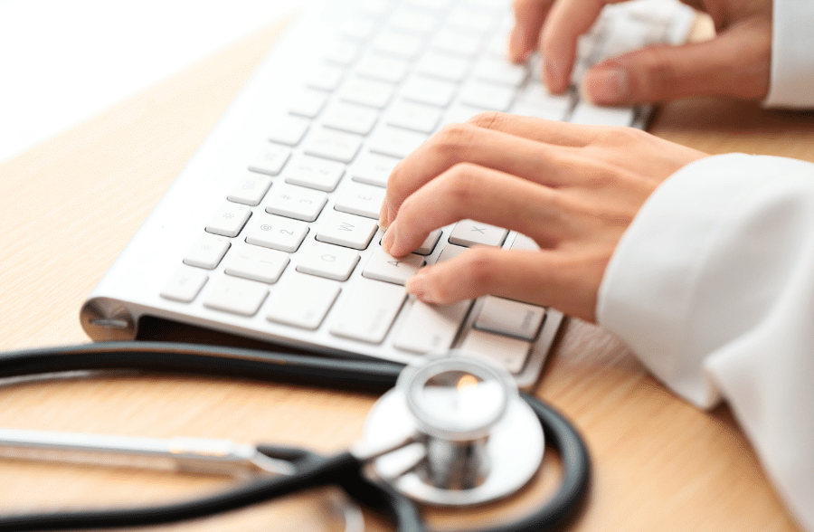 Closeup of a doctor's hands typing on a keyboard, with a stethoscope laying desk beside it.