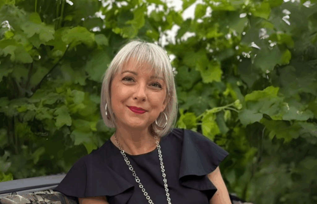 Alessandra Tolaccia smiles and sits on a bench outside in front of greenery.